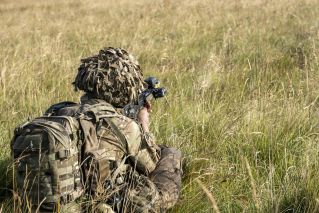 Soldier in camouflage gear aiming a rifle while crouching in a grassy field. Ideal for military themes.