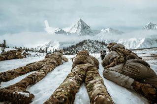 Soldiers in camouflage lying in snow during winter training in mountain landscape.